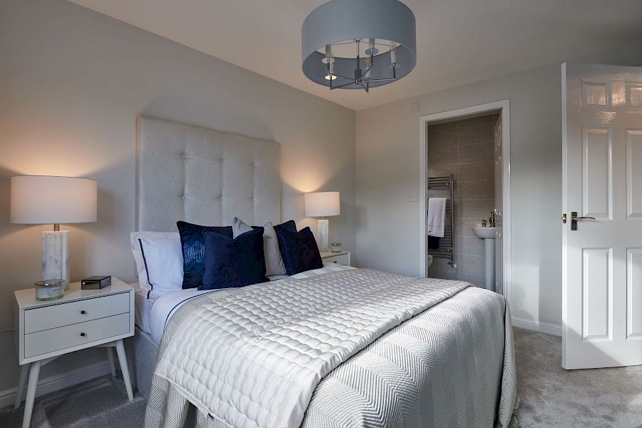Modern grey and white bedroom with double bed, navy blue cushions, en-suite bathroom, side table, white lamp and light grey carpet.